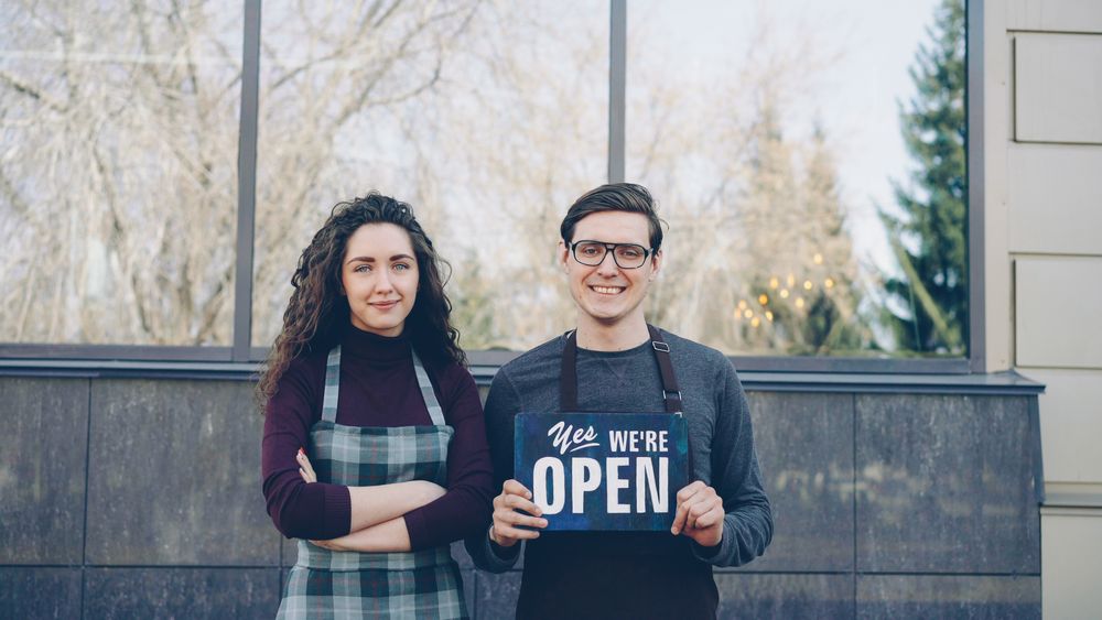 Two small business owners in aprons holding an open sign in front of their shop